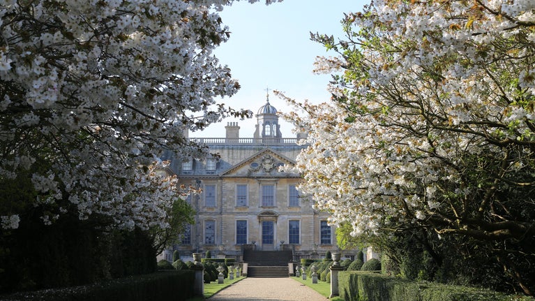 A view between two flowering blossom trees down a sand coloured path towards a large Georgian house